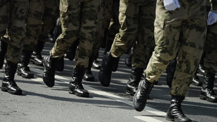Close-up view of uniformed soldiers in camouflage marching in tight formation on a street, black combat boots striking asphalt in synchronized steps during a military parade