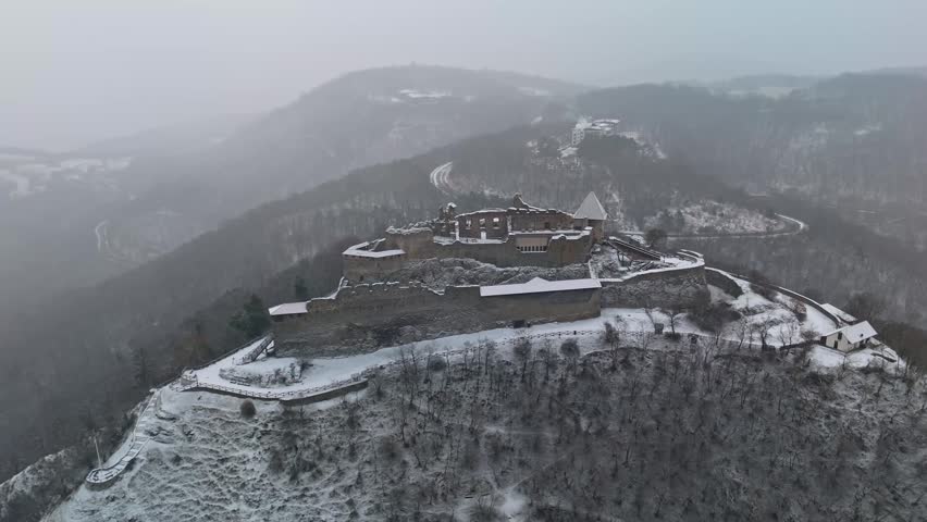 In Visegrád, Hungary, the Fellegvár castle ruins sit on a snowy hilltop amid the Visegrád Mountains and surrounding forest, viewed in winter from a drone perspective.