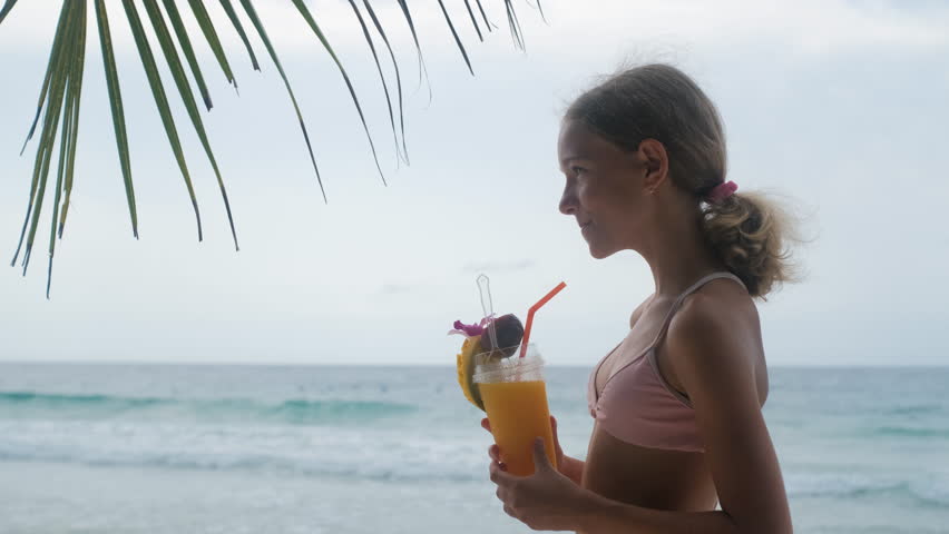 Teenage girl drinking a fresh mango smoothie through a straw on a sunny tropical beach with calm sea and sandy shore in the background. Summer refreshment concept