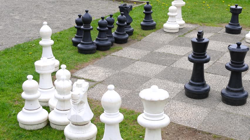 Huge chess set with large black and white pieces on a stone chessboard surrounded by green grass.