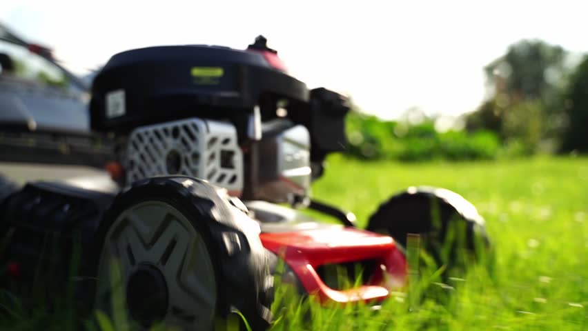 Red lawnmower sits on green grass. Sunlight glows behind blurred trees. Mower engine gleams in close-up shot. Grass blades frame foreground sharply. Perfect for garden or outdoor maintenance themes