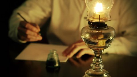 A man lit from the light of an oil lamp dips his pen in an inkwell and begins writing a letter. Focus is on oil lamp.