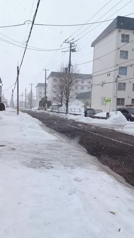 Snow falls over a peaceful city street lined with buildings, roads, and utility poles. Winter weather creates a calm atmosphere with fresh snow covering the landscape.