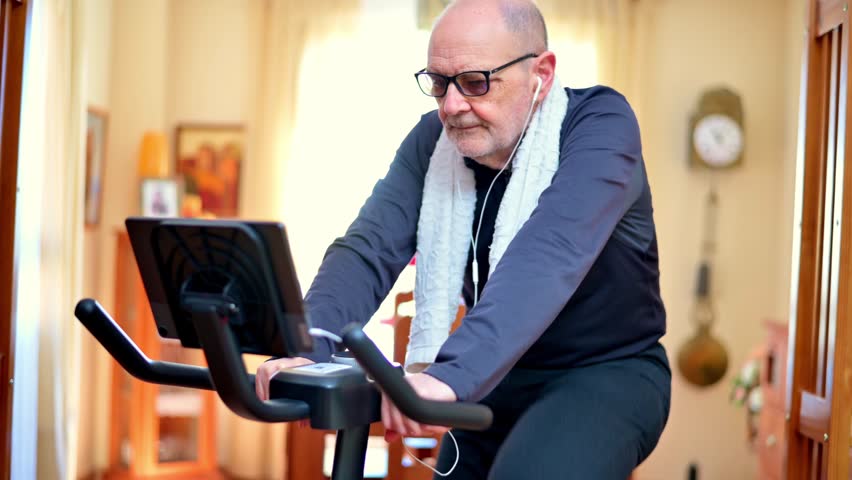 A senior man pedals a stationary bike in his living room, wearing headphones and a towel, concentrating on low-impact fitness and healthy aging with a focused, steady workout routine at home.