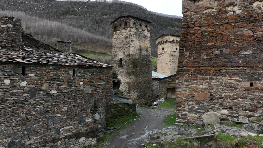 Aerial view of the ancient high altitude settlement in Ushguli, showing traditional fortified tower houses, a rushing river fork, and green valley slopes under a dramatic Caucasus mountain ridge.