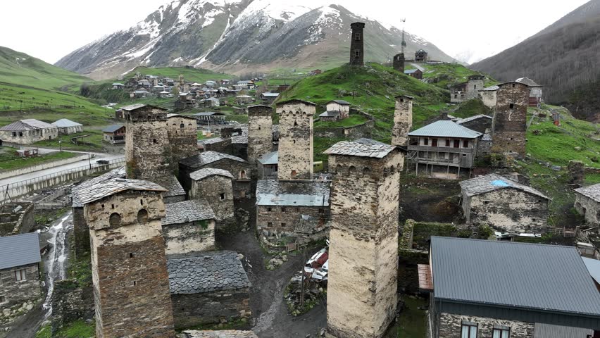 Aerial view of the ancient high altitude settlement in Ushguli, showing traditional fortified tower houses, a rushing river fork, and green valley slopes under a dramatic Caucasus mountain ridge.