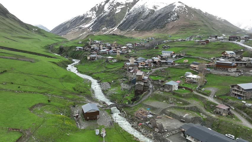 Aerial view of the ancient high altitude settlement in Ushguli, showing traditional fortified tower houses, a rushing river fork, and green valley slopes under a dramatic Caucasus mountain ridge.
