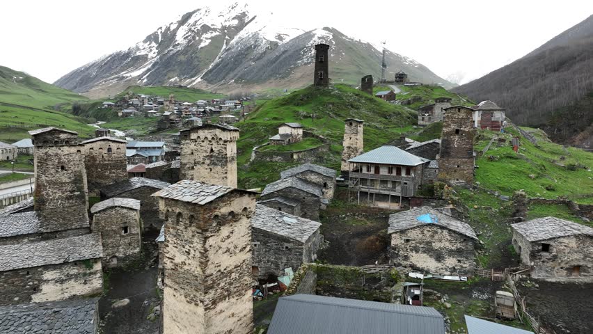 Aerial view of the ancient high altitude settlement in Ushguli, showing traditional fortified tower houses, a rushing river fork, and green valley slopes under a dramatic Caucasus mountain ridge.