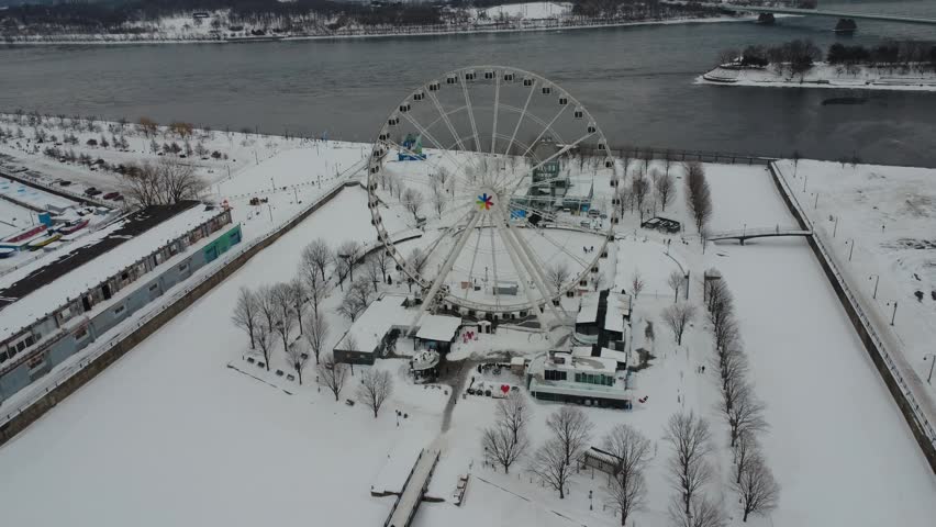 Aerial winter scene at Old Port of Montreal Canada featuring Montreal Observation Wheel, snow covered park and Saint Lawrence River beneath gray sky.