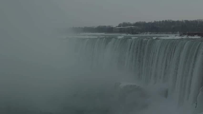 Dramatic winter view of Niagara Falls in Ontario Canada featuring partially frozen waterfall, dense mist, icy water flow and powerful natural landscape beneath gray sky