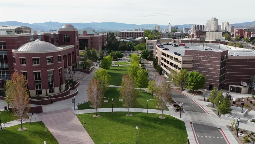 University of Nevada Reno red brick rotunda and tree-lined quad with parking structures, downtown Reno skyline and arid mountains beyond under cloudy and sunny sky. Nevada, USA. Aerial ascend shot.