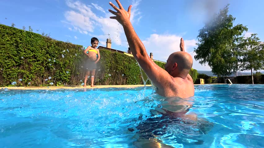 Playful son with arm floaties bravely jumping into the clear blue water of a swimming pool, being caught by his smiling father during a beautiful sunny summer day creating a splash