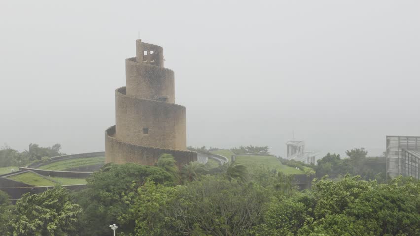 MOTOBU, OKINAWA, JAPAN – JUNE 2, 2026: A video shows the brick-red Observation Tower at the Tropical Dream Center heavily isolated against a bleak, storm-swept sky as Typhoon Jangmi (Typhoon No. 6) barrels directly over Okinawa. Driven by the powerful storm system, thick grey clouds roll heavily across the frame, completely blanketing the background so that the East China Sea is entirely invisible. Intense wind fields and torrential downpours lash the structure continuously, creating a dense, shifting haze that blurs the surrounding landscape. The nearby tropical foliage and palm trees whip violently in the frame, buffeted by fierce gusts of up to 40 meters per second as the core of the typhoon makes its closest approach, completely obscuring the sweeping panoramic vistas normally seen from the park.