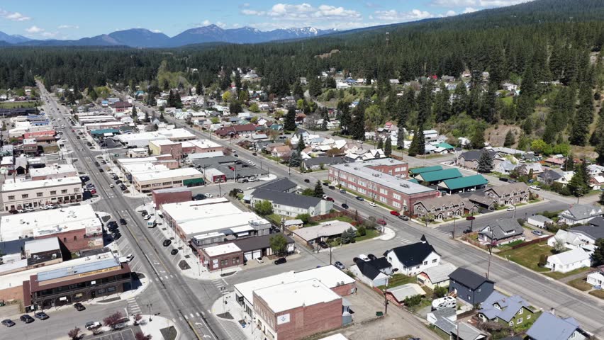 Small town main street with mix of commercial and residential buildings, evergreen forest and snow-capped mountain peaks rising beyond under sunny blue sky. Washington State, USA. Aerial flyover.