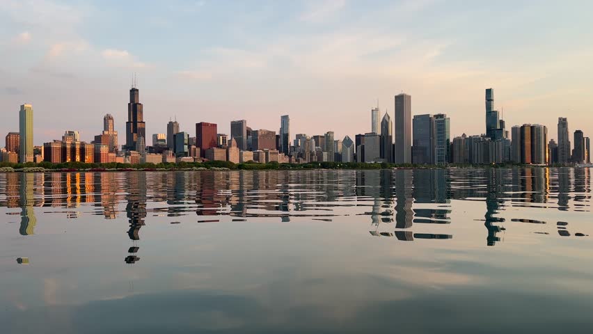 Chicago skyline panorama viewed from Lake Michigan. Skyscrapers mirror in calm water. Sunrise light casts warm glow. Cityscape aerial footage.