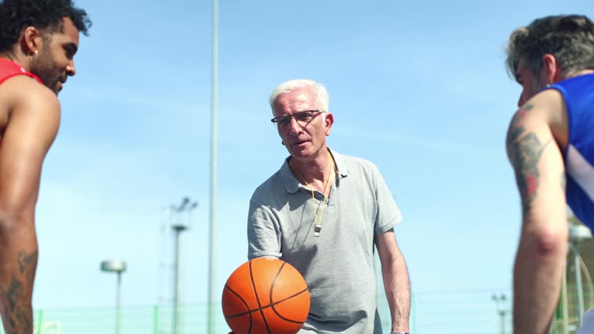 Senior referee throwing a basketball in the air to start a game between two opponents on an outdoor court under a clear blue sky