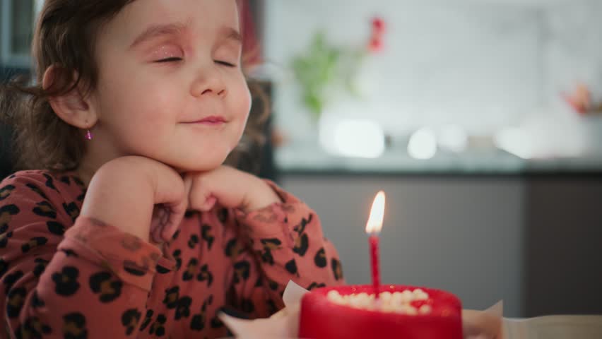 Little girl blows out a birthday candle. She closes her eyes, focused and calm. Her cheeks puff as she breathes out. Her hands rest under her chin, still and patient. She wears a patterned top, soft