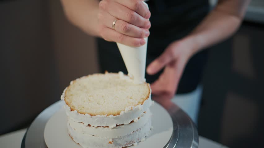 Close-up of a person decorating a cake with white frosting. Hand squeezes piping bag steadily, focusing on cake