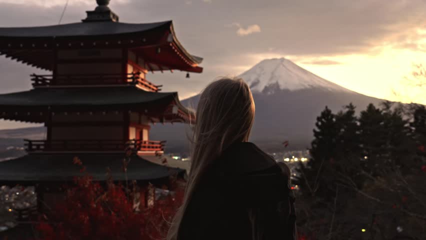 Back view of a blonde woman standing at Arakura Sengen shrine looking at the iconic Chureito Pagoda and Mount Fuji with autumn leaves and city lights at sunset in Japan