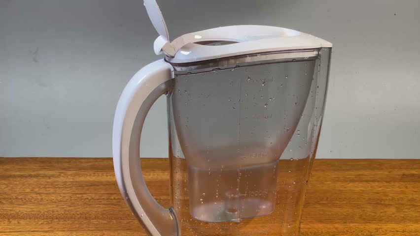 Pouring cold water into empty plastic transparent tap water purifier jug standing on a wooden table, view close-up
