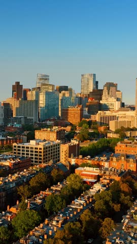 Cinematic drone shot flying over residential neighborhoods toward downtown skyline at golden hour. Boston urban landscape with historic brownstones and modern glass office towers in Massachusetts.
