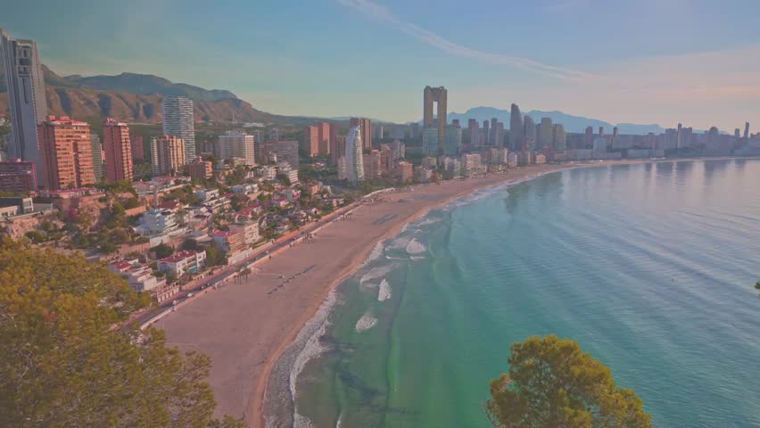 Visitors walk along the sandy beach in Benidorm while others enjoy the water. Sunny skies surround the tall buildings along the coast during midday
