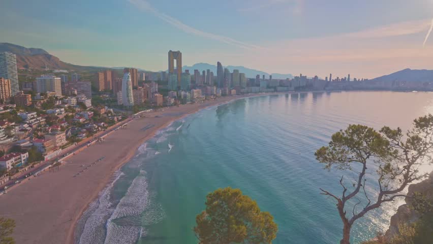 The coastline of Benidorm with clear water, sandy beach, and the city skyline in the background. It captures a bright afternoon, highlighting the beauty of this popular location