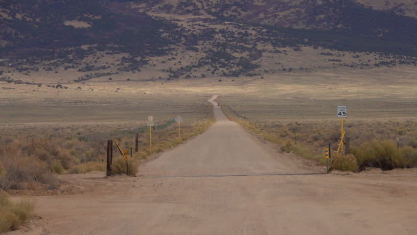 VERTIGO ZOOM EFFECT POV: Driving along empty dirt road towards Colorado Rockies in United States. Straight gravel road leading towards Rocky Mountains, black raven bird flying across the dusty route
