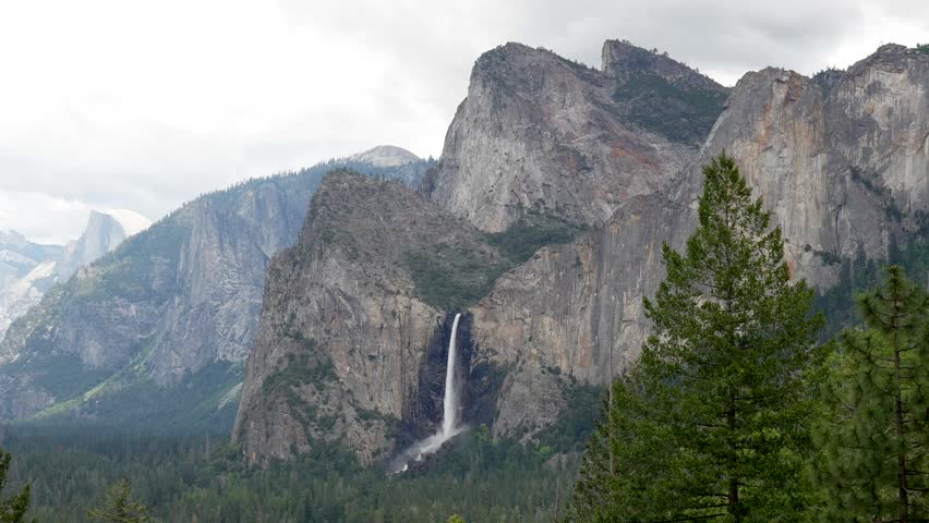 Majestic view from tunnel view showing bridalveil fall cascading down granite cliffs into the lush yosemite valley, with the iconic half dome visible in the distant background on a cloudy day