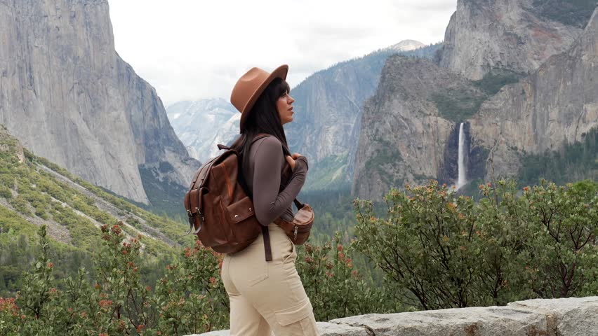 Happy female hiker with a backpack standing at tunnel view overlook, admiring the stunning landscape of yosemite valley with bridalveil falls and el capitan in the background