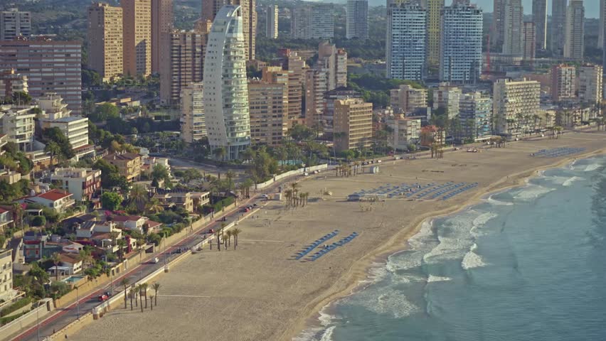 Benidorm shows its sandy beach with people walking along the shore and relaxing under umbrellas. The modern buildings rise above the sandy coast. It is a bright and sunny day