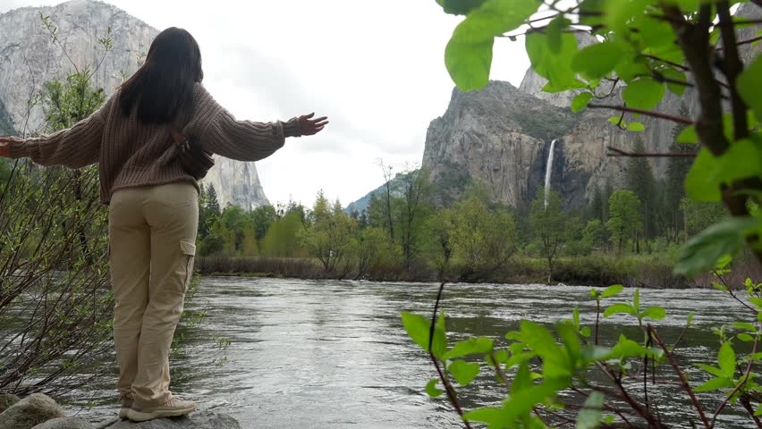 Young woman standing on a rock with arms outstretched, feeling freedom while admiring the breathtaking landscape of yosemite valley, with bridalveil fall and el capitan in the background