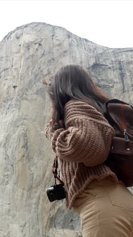 Young smiling photographer with a backpack looking up at the stunning granite monolith of el capitan, her hair blowing in the wind during a hike in yosemite national park, california