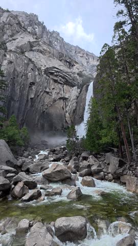 Lower yosemite falls majestically cascades down granite cliffs, feeding a rocky stream surrounded by evergreen trees in california