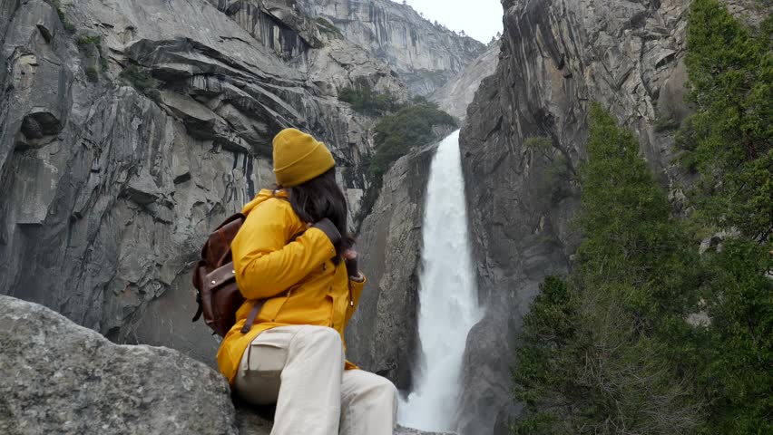 Happy female hiker in a bright yellow jacket sitting on a rock, admiring the stunning view of the powerful lower yosemite falls and looking at the camera, surrounded by granite cliffs