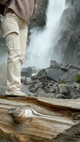 Young woman in a yellow beanie and backpack stands on a log, gazing at the powerful lower yosemite falls, immersed in the forested valley and wild sierra nevada landscape