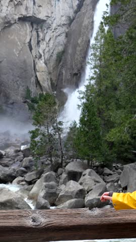 Young female traveler in a yellow raincoat enjoying the breathtaking view of lower yosemite falls, leaning on a wooden railing and smiling at the camera in the middle of a forest in california