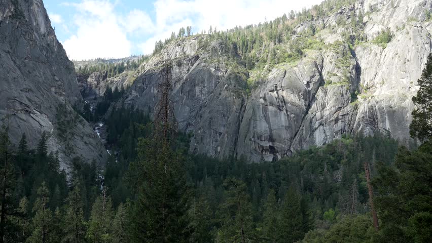 Majestic granite mountains towering over a lush pine forest on a beautiful sunny day, with a glimpse of a cascading waterfall along the famous mist trail in yosemite national park, california