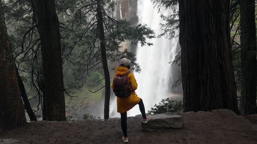 Young woman hiker wearing a yellow raincoat and carrying a backpack admiring the powerful vernal fall waterfall while trekking along the mist trail in yosemite national park, california