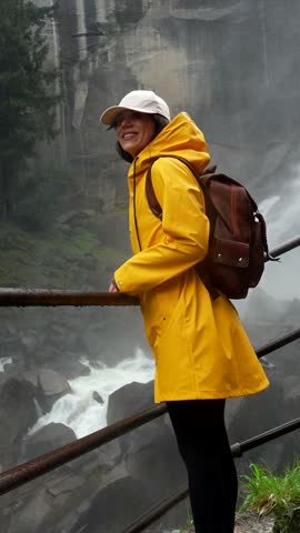 Young smiling woman in a yellow raincoat hiking on the wet, misty stone steps of the mist trail, enjoying the spectacular view of vernal fall in yosemite national park, california