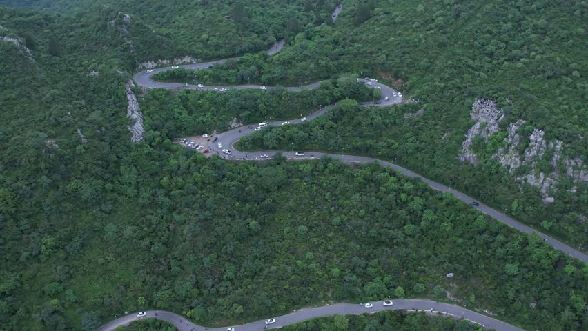Drone Aerial of Winding Switchback Mountain Road with Rocky Peaks and Forest Valley. Aerial drone footage of multiple switchback roads snaking through a lush forested mountain valley with rugged rocky limestone peaks emerging from the green canopy on both sides. A busy scenic viewpoint with gathered visitors and parked vehicles is visible mid-frame. Ideal for travel documentaries, geography features, and South Asian mountain content. Filmed in Margalla Hills, Islamabad, Pakistan.