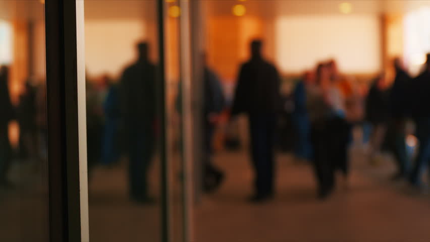 Blurred crowd of people walking through modern lobby entrance with warm light and reflections, 4K selective focus