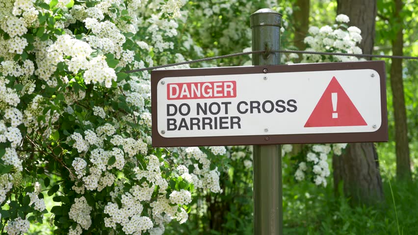 Warning sign on a garden barrier beside blooming white shrubs in a wooded outdoor setting