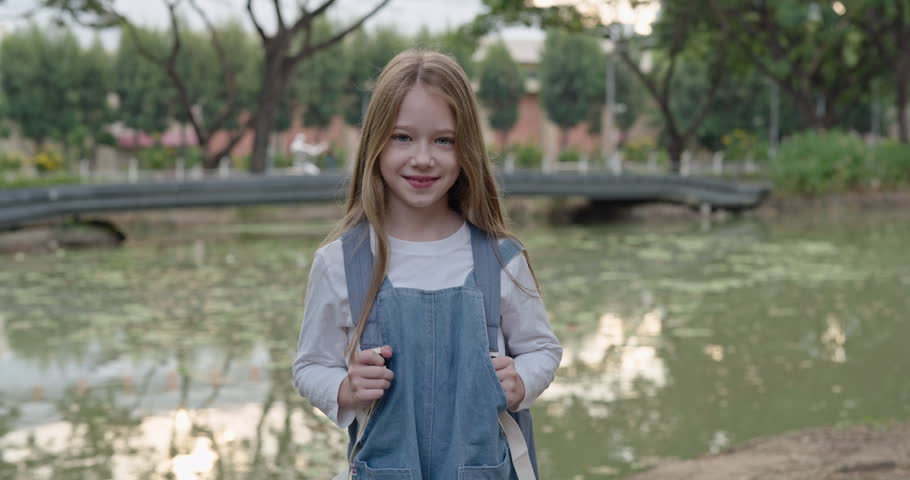 Portrait of happy little schoolgirl with backpack stands near pond. Adorable young girl with schoolbag smiles in green urban park. Positive elementary student