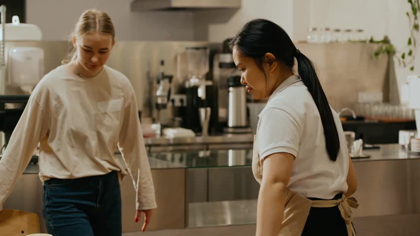 A female intern and a female barista are sitting at a table holding cups while talking and laughing