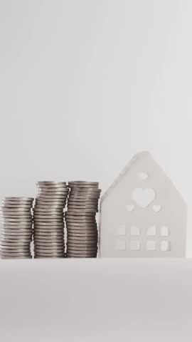 The hand moves into the frame on the left and places coins in the foot next to a model of a white ceramic house on a white surface. The stack grows as the hand removes the coins one by one.