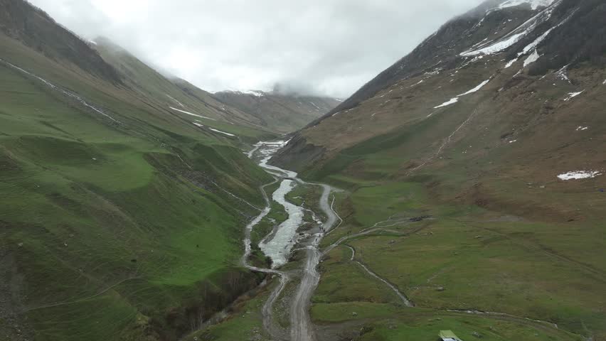 High-angle aerial view capturing a clean, rushing mountain creek flowing from a melting snow patch down a vibrant green alpine valley with rocky terrain and small wildflowers in Ushguli, Georgia
