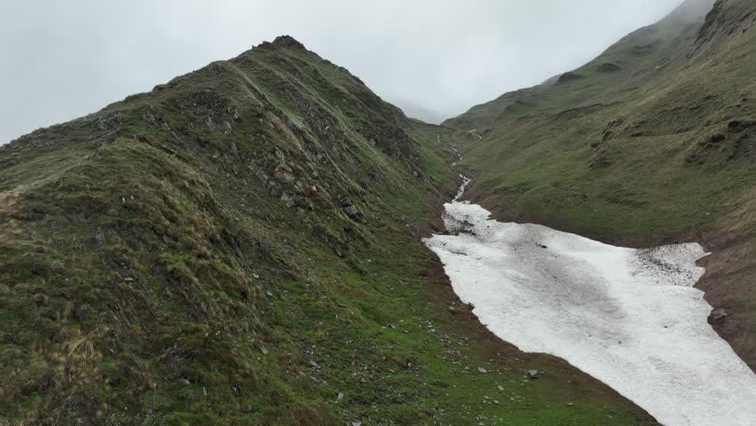 High-angle aerial view capturing a clean, rushing mountain creek flowing from a melting snow patch down a vibrant green alpine valley with rocky terrain and small wildflowers in Ushguli, Georgia