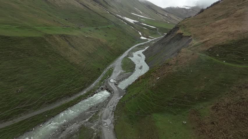 High-angle aerial view capturing a clean, rushing mountain creek flowing from a melting snow patch down a vibrant green alpine valley with rocky terrain and small wildflowers in Ushguli, Georgia