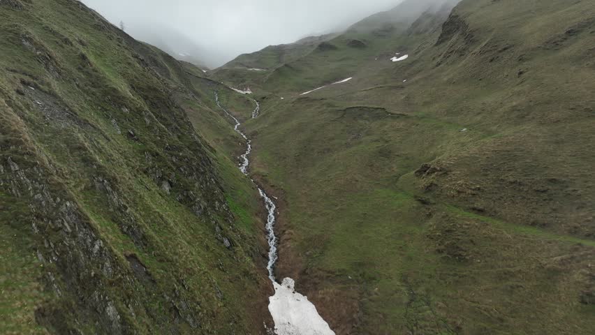 High-angle aerial view capturing a clean, rushing mountain creek flowing from a melting snow patch down a vibrant green alpine valley with rocky terrain and small wildflowers in Ushguli, Georgia